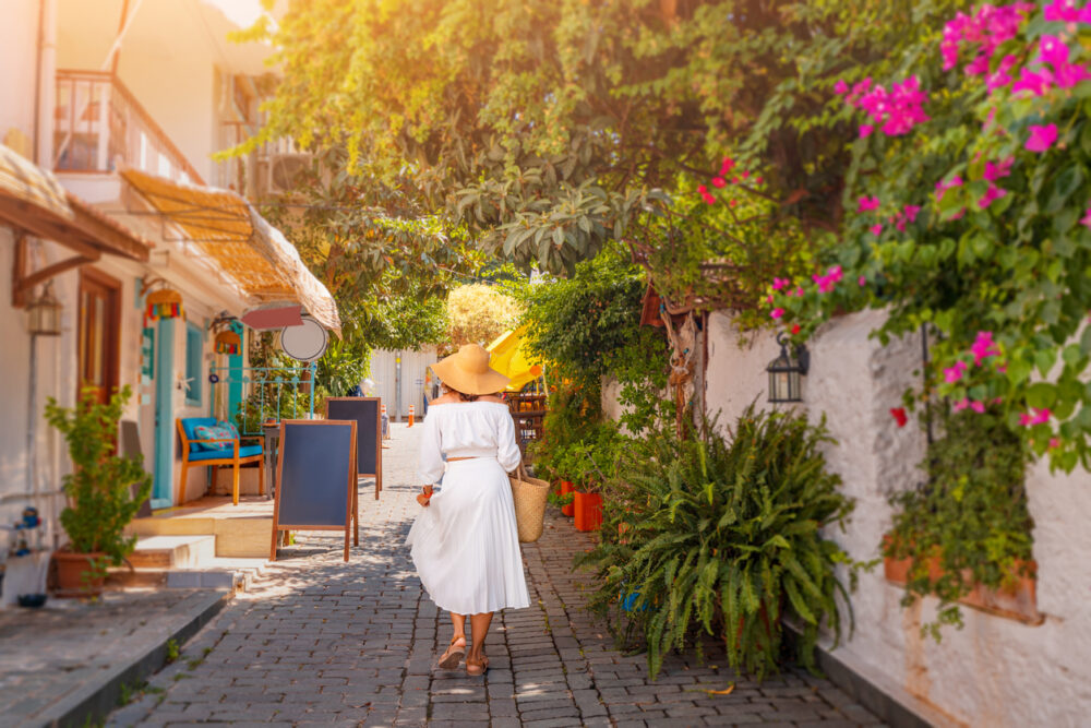 Woman in white dress and hat walks down a cobblestone street lined with greenery and shops.
