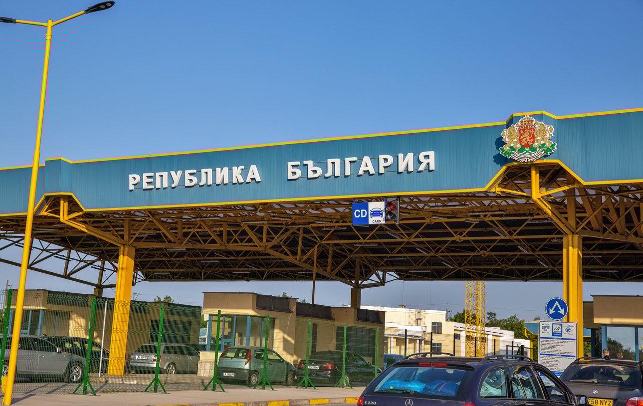Border checkpoint with "Republika Bulgaria" sign, cars waiting under a large canopy structure.