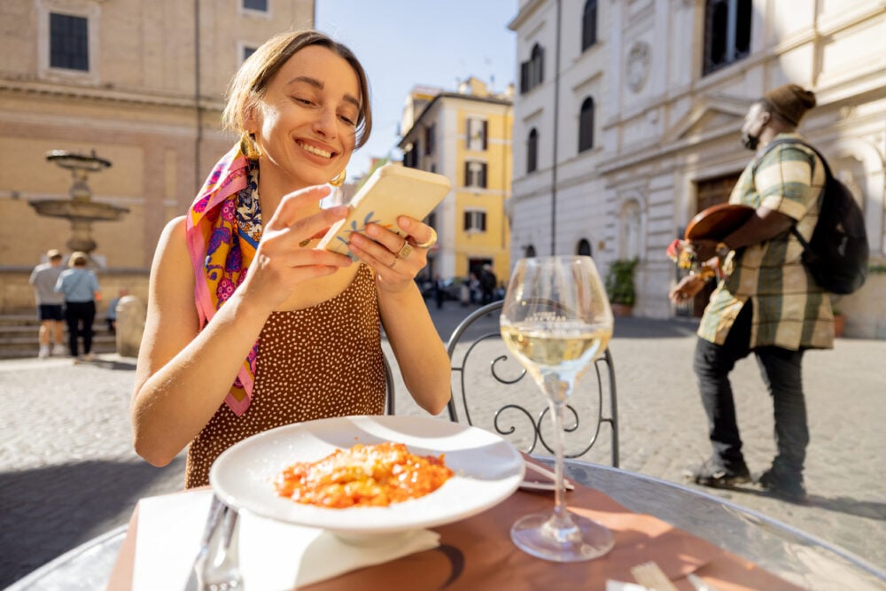 Woman smiling at smartphone, seated at outdoor cafe with pasta dish and wine, in sunny square.