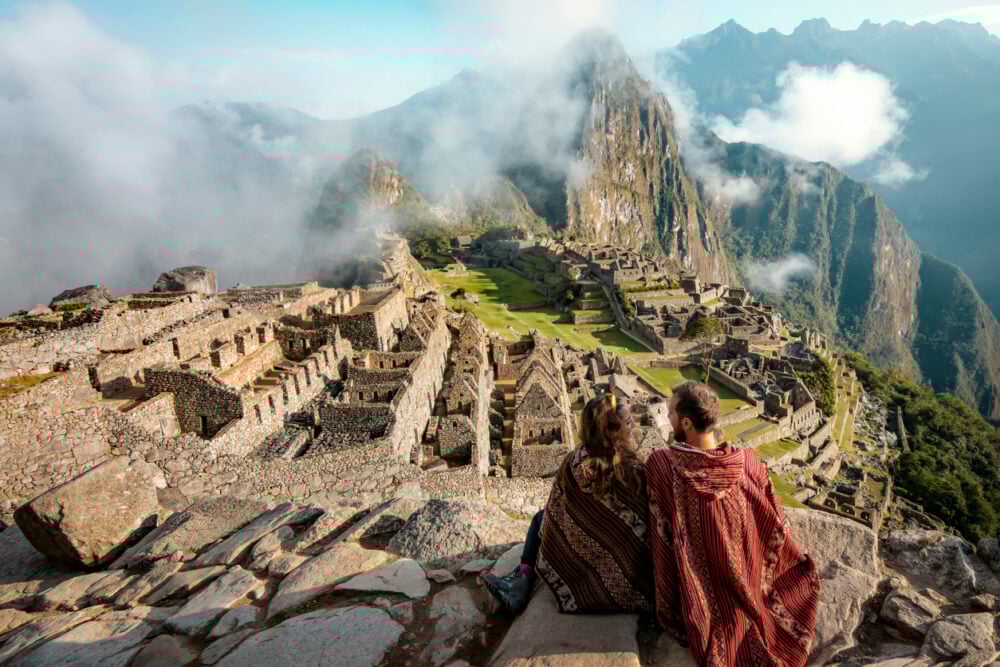 Two people in traditional clothing sit overlooking the ancient ruins of Machu Picchu surrounded by mountains and mist.
