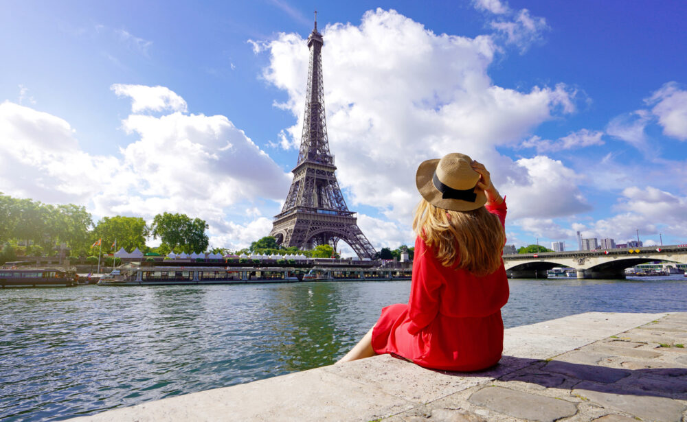 Woman in red dress sitting by river, facing Eiffel Tower, under blue sky with clouds.