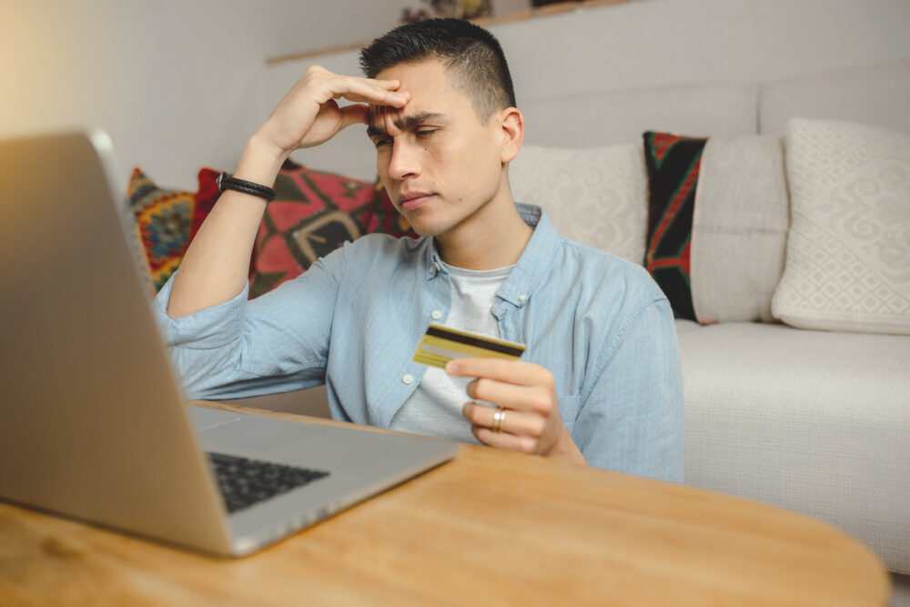 Man holding credit card, looking at laptop with a worried expression, sitting on a couch.