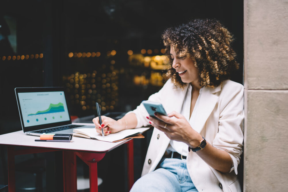 Woman writing in notebook, holding phone, with open laptop showing graphs on table.