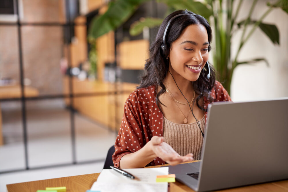 Woman wearing headphones, smiling at laptop, with a notebook and pen on a desk in an office.