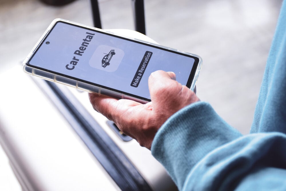 Person holding a smartphone with "Car Rental" and "Make a Reservation" on the screen, next to a suitcase.