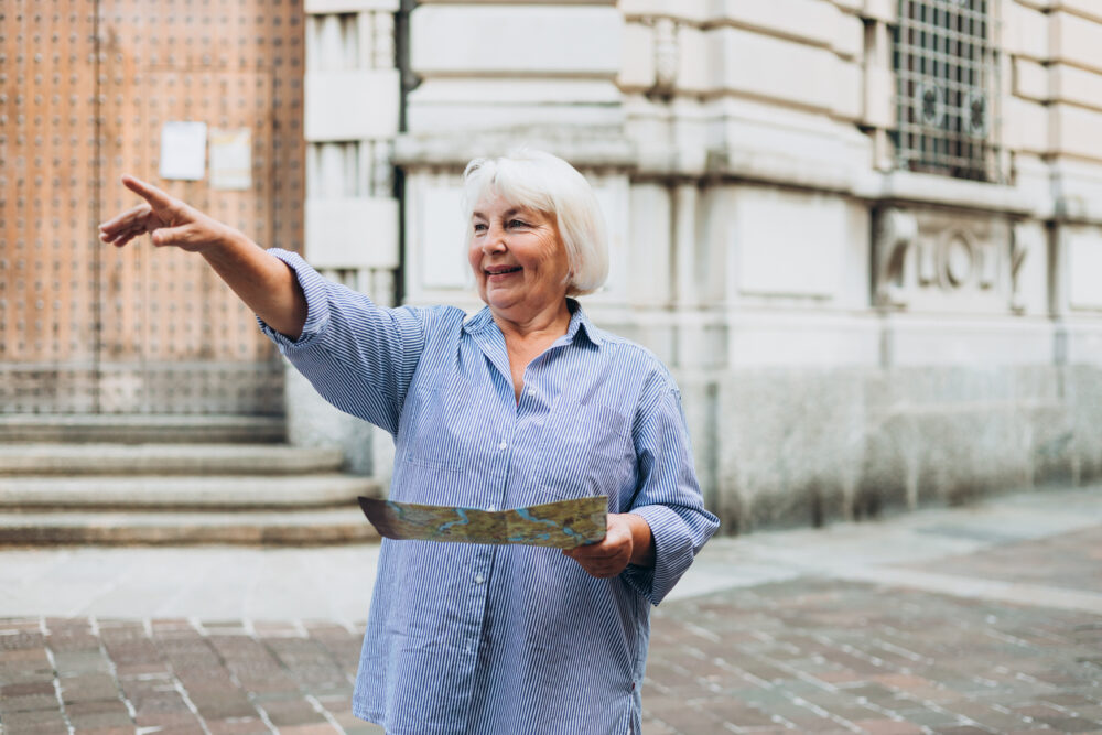 Elderly woman holding a map and pointing, standing outside a historic building.