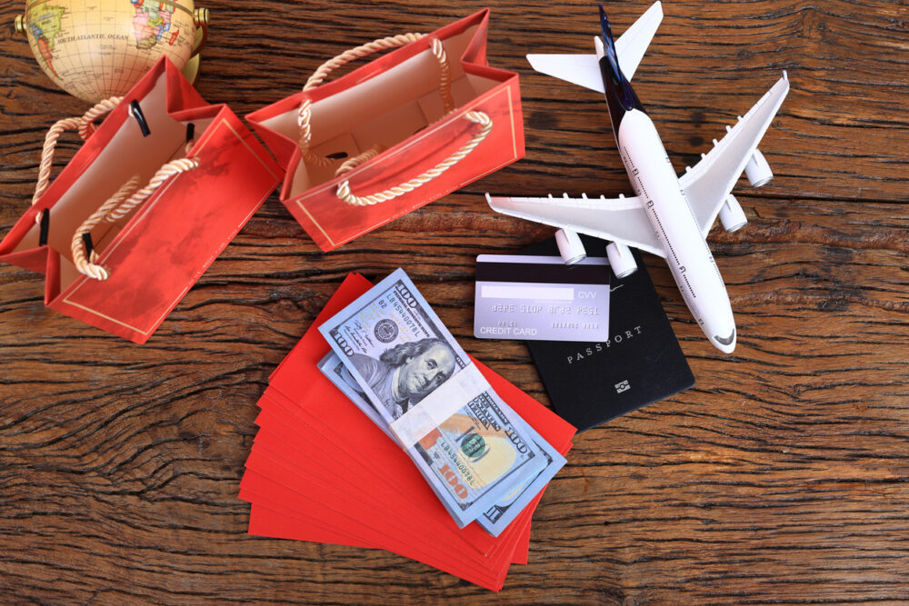Model airplane, red gift bags, globe, cash, passport, and credit card on wooden surface.