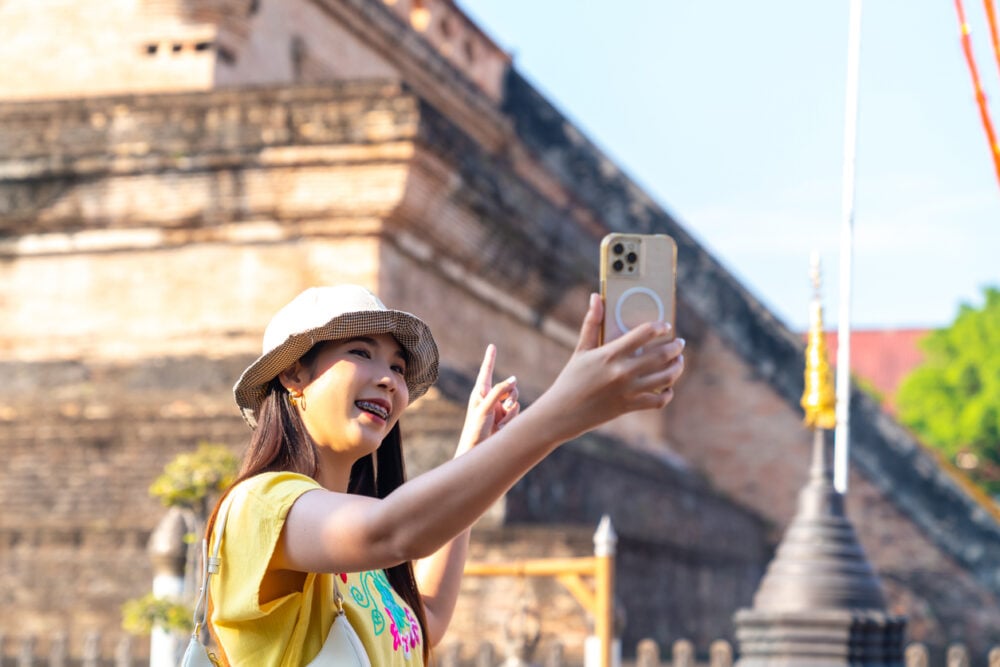 Young woman taking a selfie with a smartphone, standing in front of an ancient brick structure.