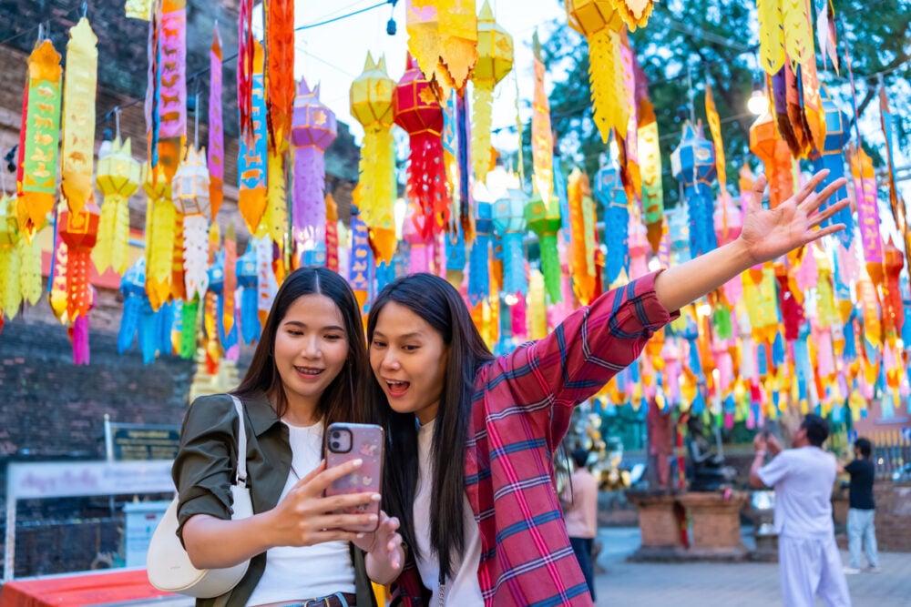 Two women take a selfie under colorful hanging lanterns at an outdoor festival.