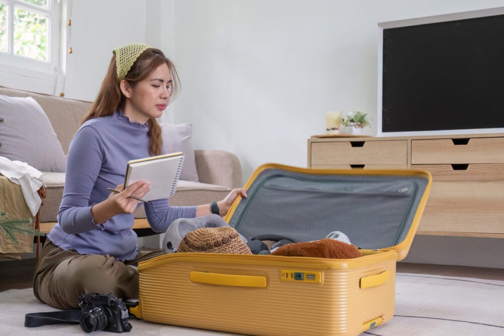 Woman sitting on floor, packing items into a yellow suitcase, holding a notepad and pen.