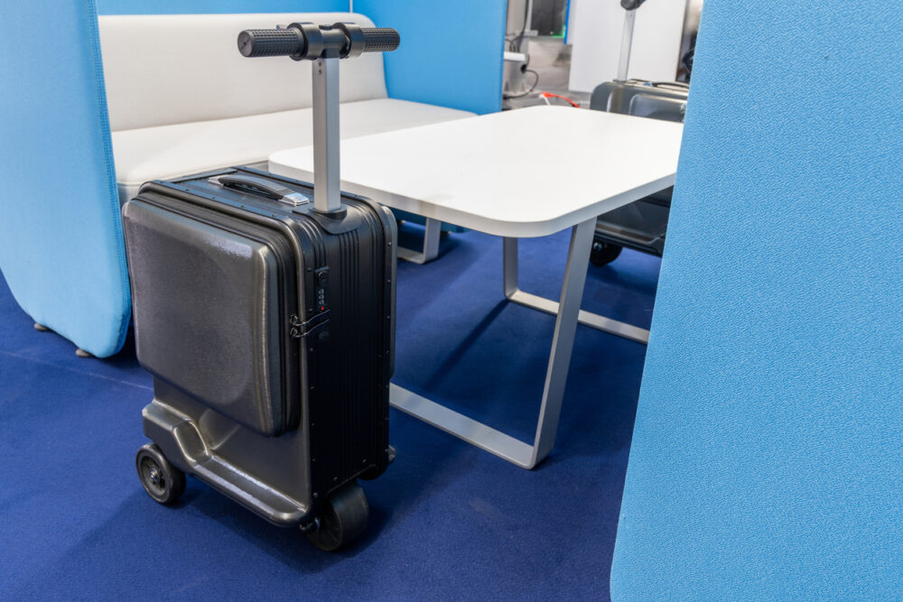 Black suitcase with integrated scooter handle, next to a table and blue partition on a carpeted floor.