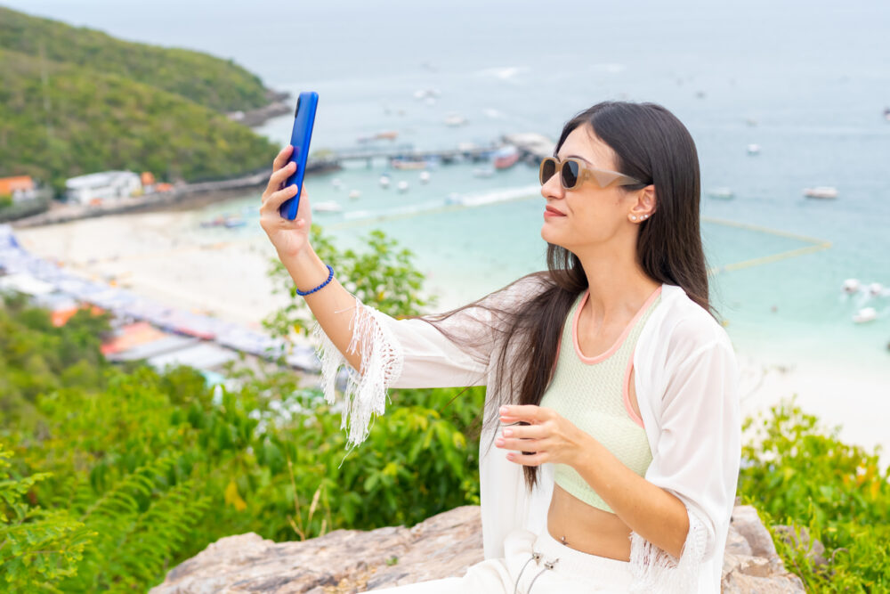 Woman taking a selfie with a blue smartphone near a beach, wearing sunglasses and a white outfit.