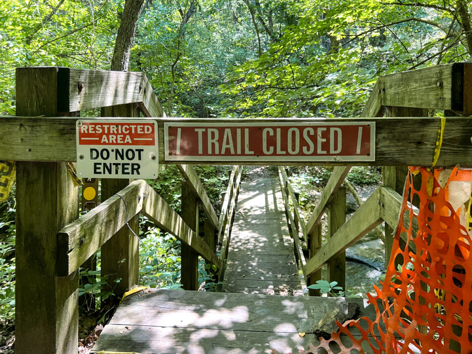 Wooden bridge with signs reading "Trail Closed" and "Restricted Area Do Not Enter." Orange barrier on right.