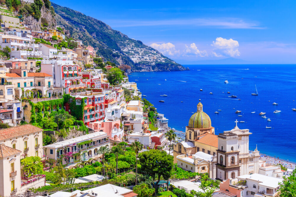 Colorful hillside buildings overlooking a blue sea with boats, lush green foliage, and a dome-shaped church.
