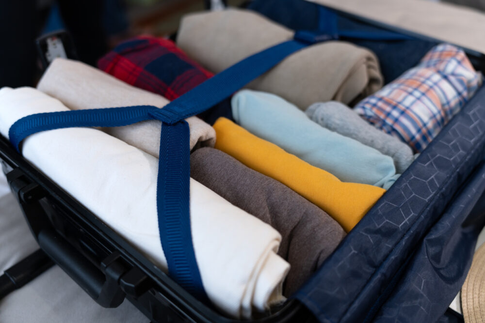 Open suitcase with neatly rolled clothes, secured by a blue strap, including shirts and trousers.