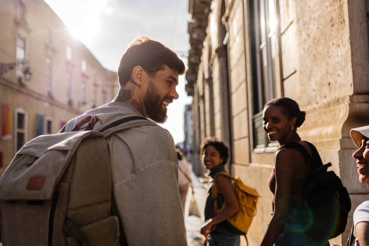 Four people with backpacks walking and laughing on a sunlit street lined with buildings.