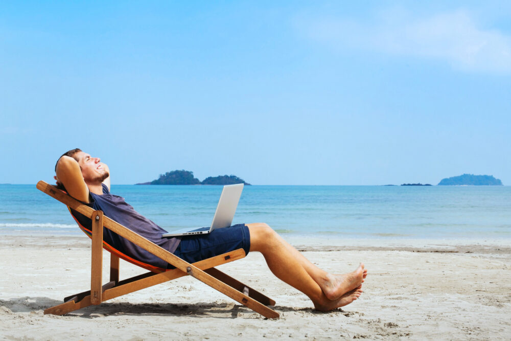 Man relaxing on beach chair, with laptop on lap, by the ocean under clear blue sky.