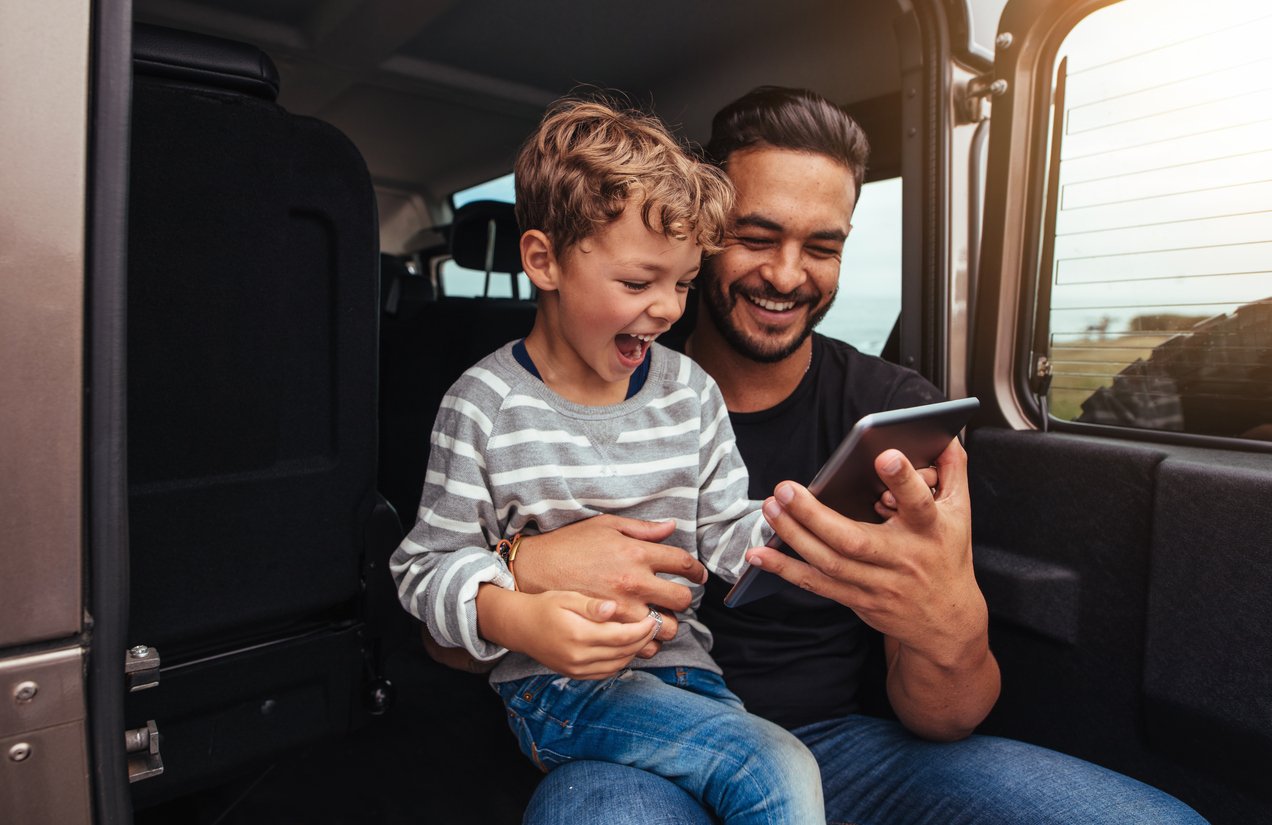 Man and child sitting in a car, smiling and looking at a tablet together.