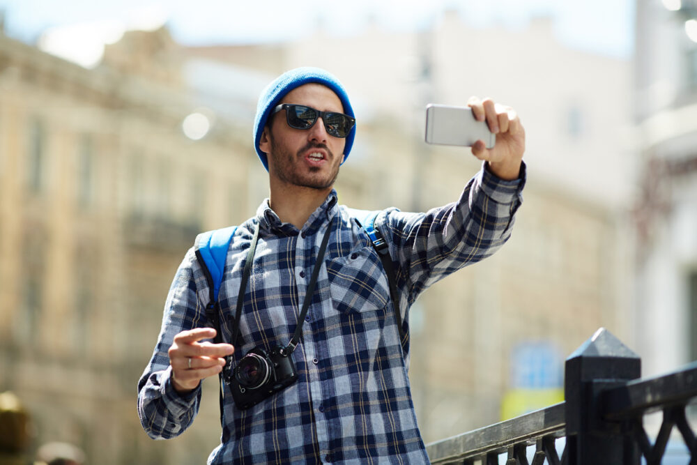 Man in blue beanie and plaid shirt taking a selfie outdoors with camera around neck.