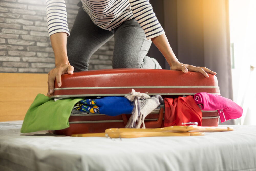 Person struggling to close overfilled red suitcase with clothes spilling out on bed.