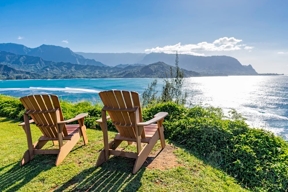 Two wooden chairs overlook a scenic coastal view with mountains and ocean under a clear blue sky.