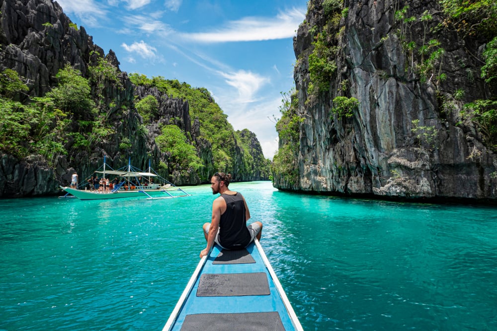 Person sitting on boat in turquoise water, surrounded by tall, rocky cliffs with green vegetation under a blue sky.