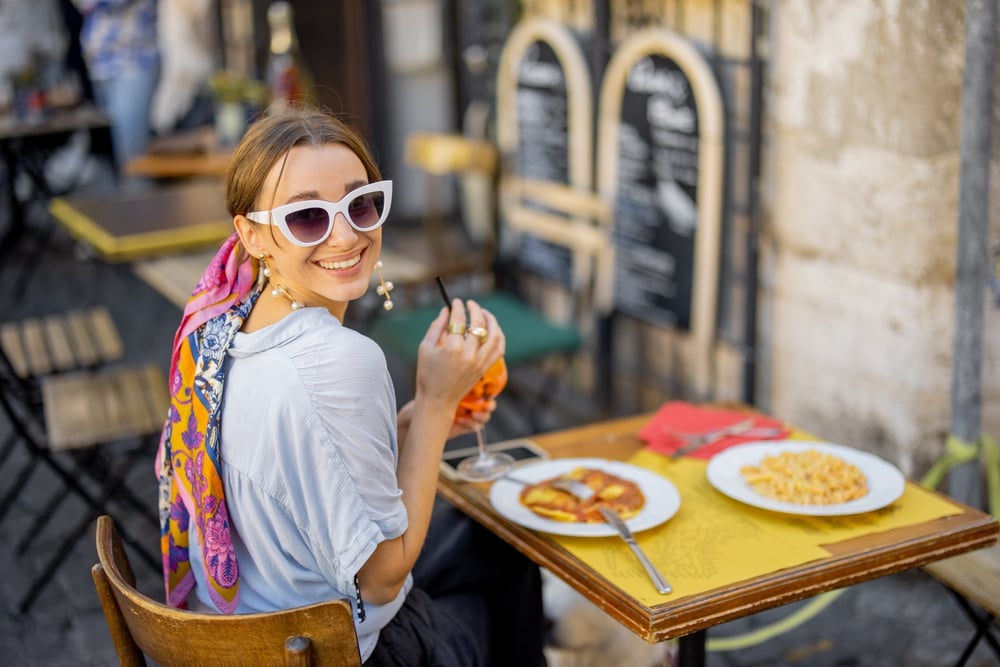 A woman in sunglasses dining outdoors, holding a drink, with pasta dishes on the table.