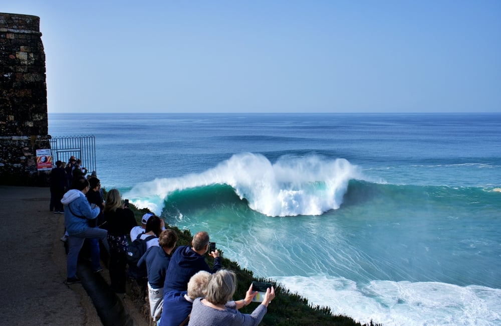 People watching and photographing large ocean wave from a cliffside path on a clear day.