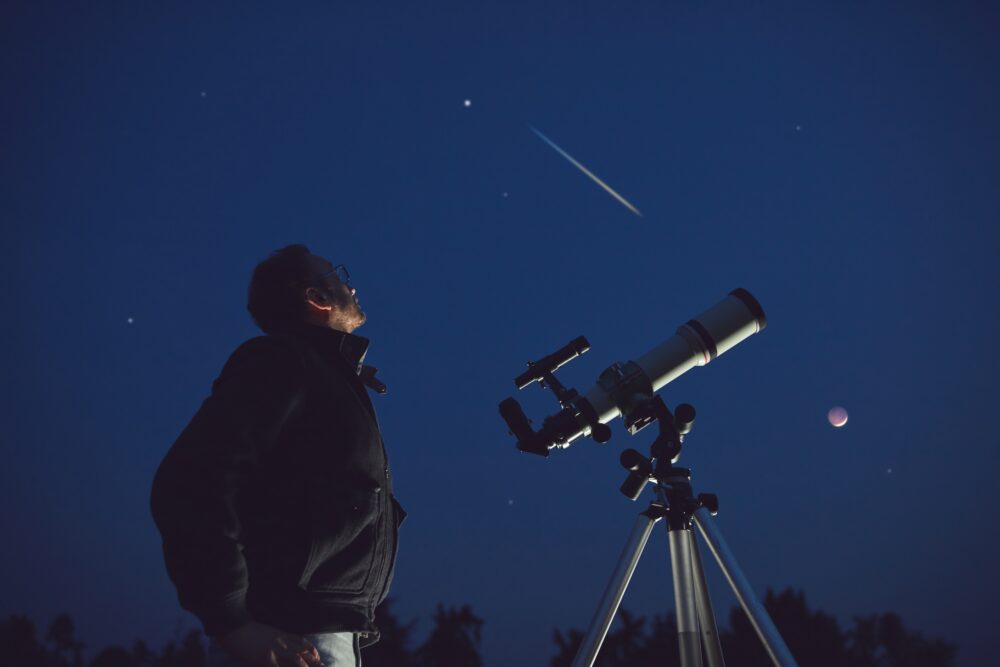 Man looking at sky beside telescope under dark, starry night, with a visible shooting star and moon.