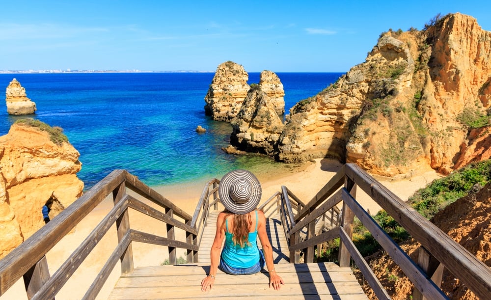 A woman with a striped hat sits on wooden stairs leading to a beach with rock formations.