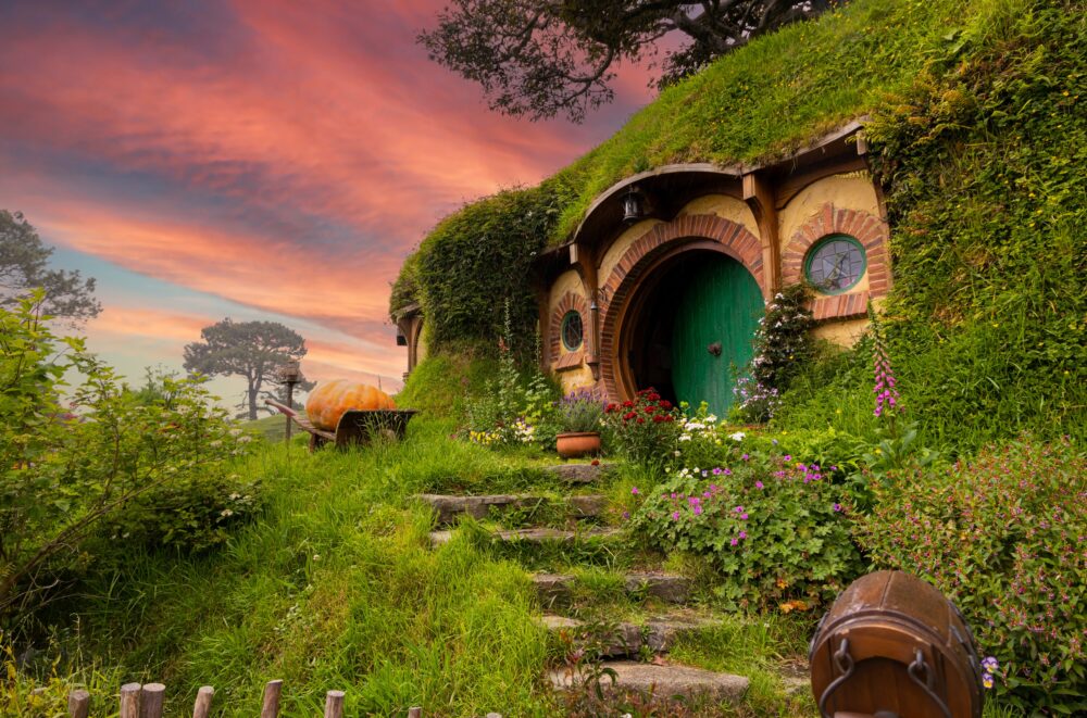 Hillside house with round door surrounded by greenery and colorful flowers, under a vibrant sunset sky.