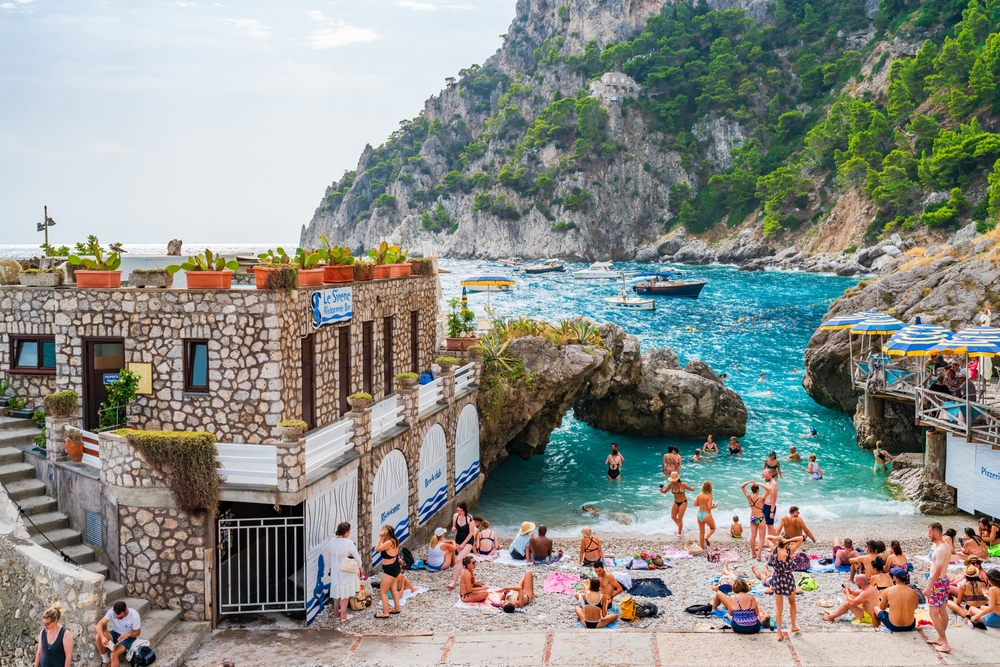 People relaxing on a rocky beach with clear blue water, boats, and a stone building.