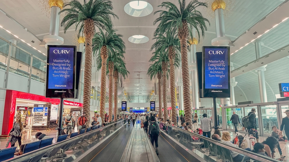 Wide airport corridor with palm trees, moving walkway, advertising screens, and people walking or seated.