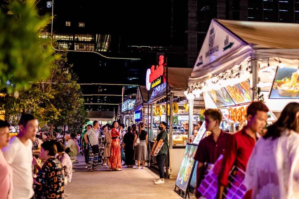 People walking and shopping at a bustling outdoor night market with food stalls, lights, and signs.