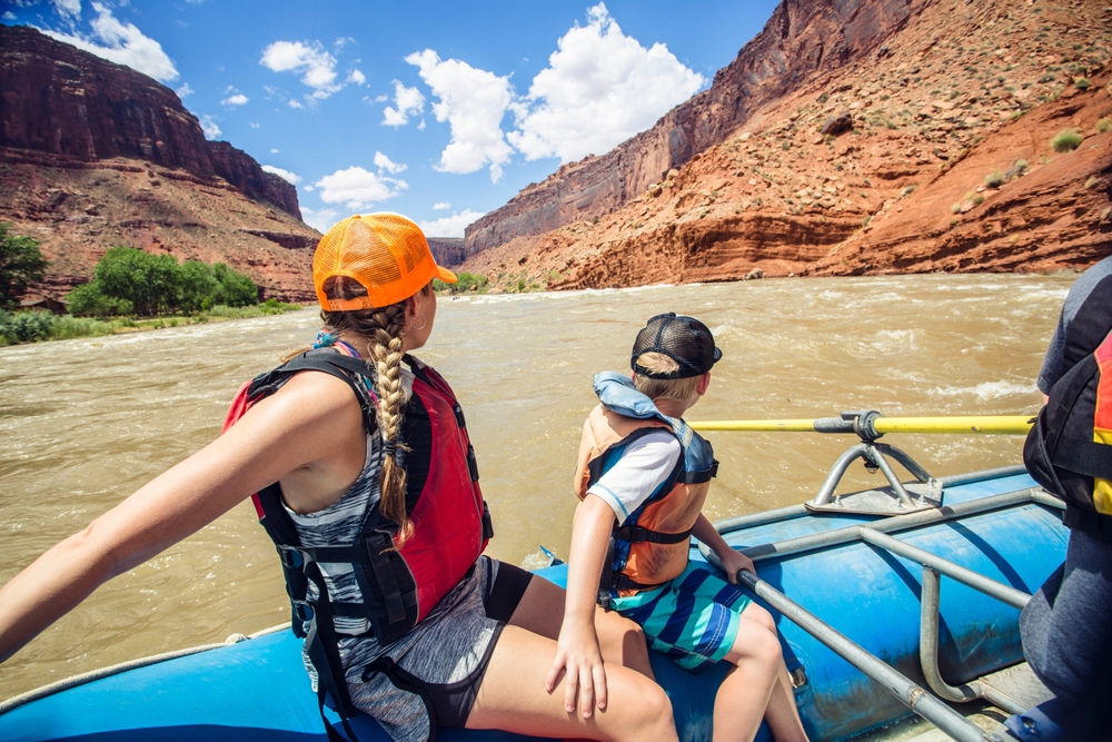 Two people rafting on a river between red rock canyons under a blue sky with clouds.