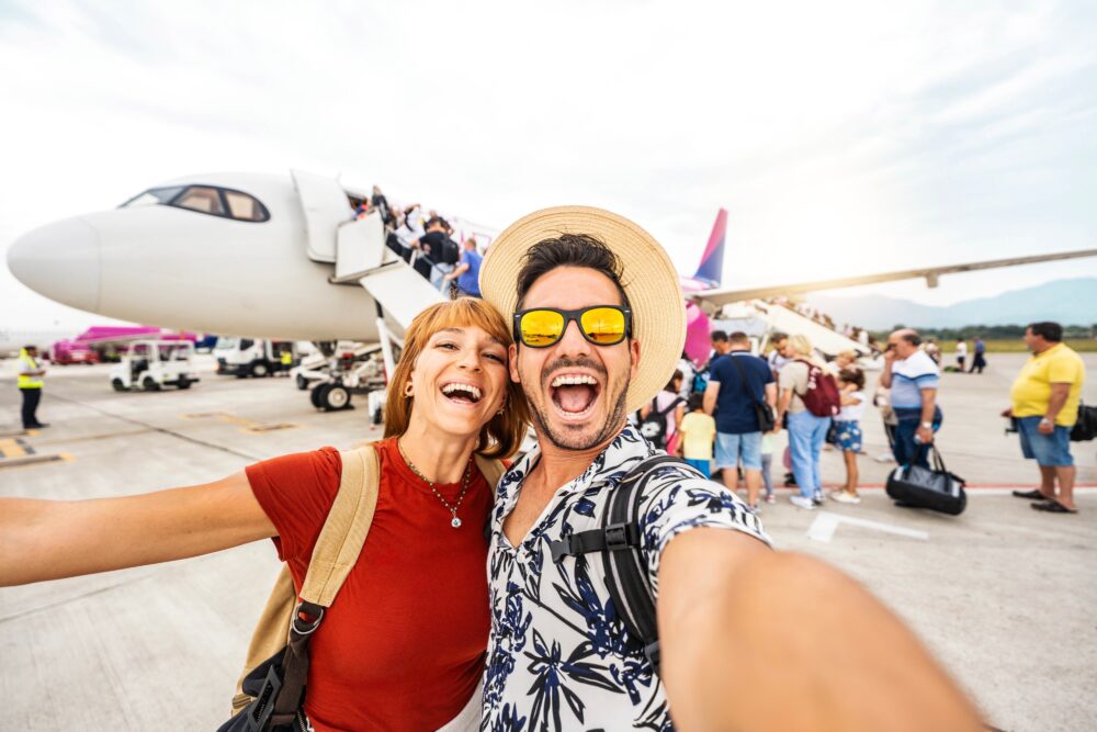 Two people taking a selfie in front of an airplane, with passengers boarding via a staircase.