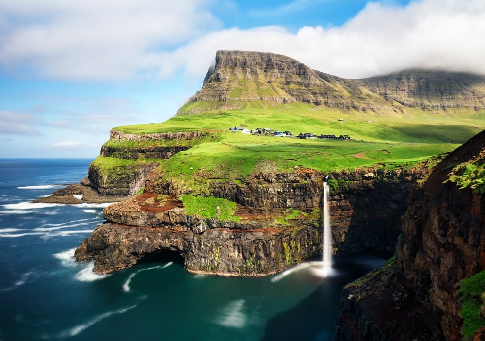 Cliffside village with green grass, waterfall, and ocean under a partly cloudy sky.