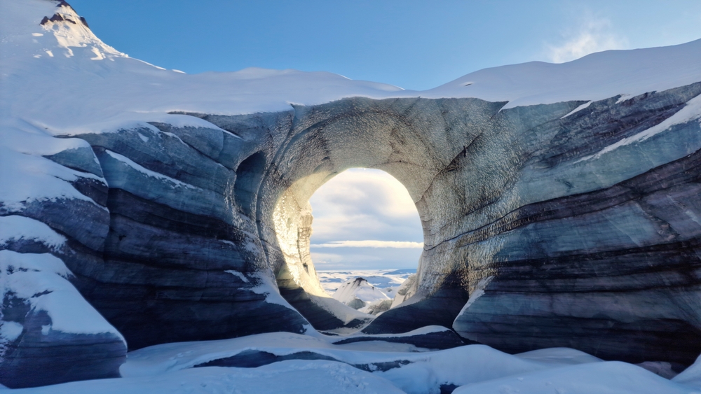 Ice cave with a circular opening, surrounded by snow and illuminated by sunlight.