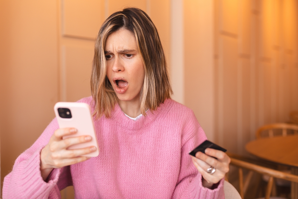 A woman in a pink sweater looks shocked at her phone while holding a credit card.