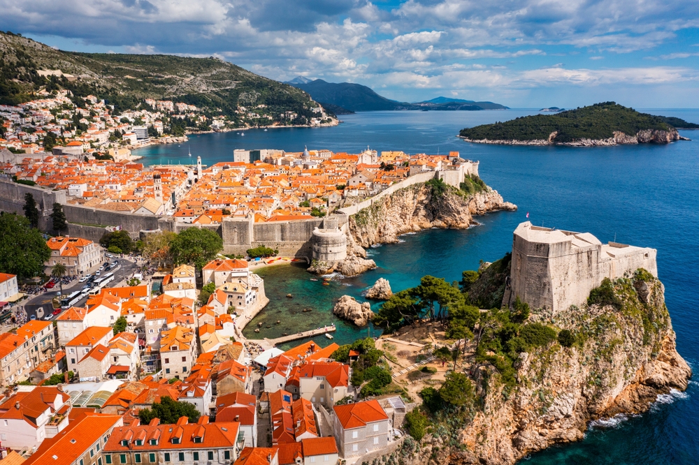 Aerial view of fortified city with orange-roofed buildings by the sea, surrounded by stone walls and cliffs.