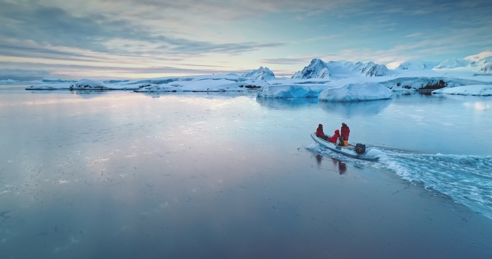 Two people in a small boat on icy waters with snow-covered mountains in the background.