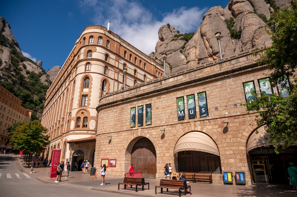 Historic stone building in front of rocky hillside, with people walking and benches along the sidewalk.