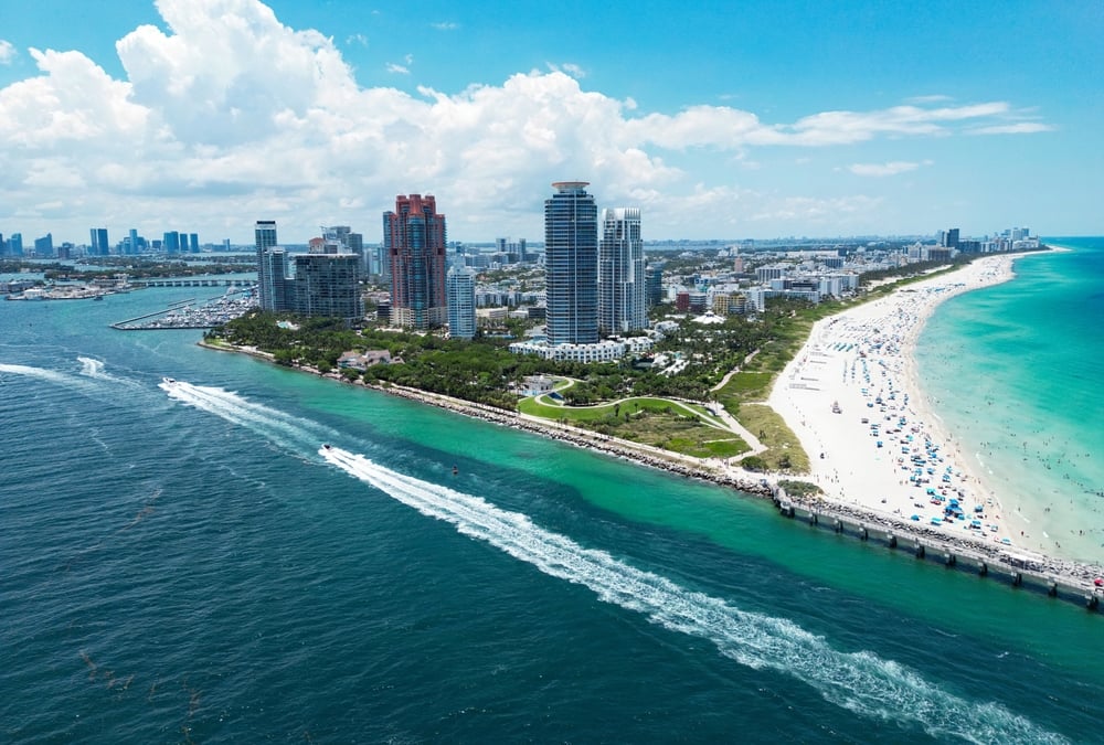 Aerial view of a beach with skyscrapers, green water, and numerous people sunbathing and swimming.