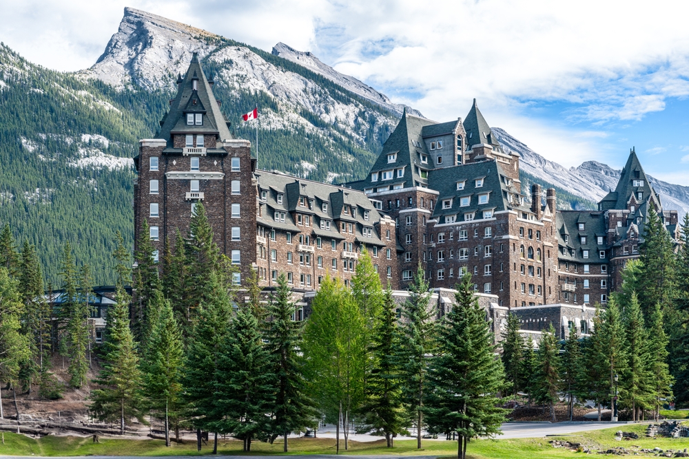 Grand castle-like hotel surrounded by forested mountains under a partly cloudy sky.