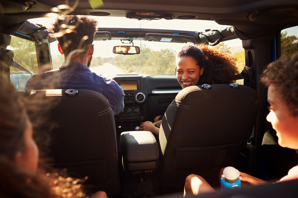 Two adults and two children smiling inside a car during a daytime drive.