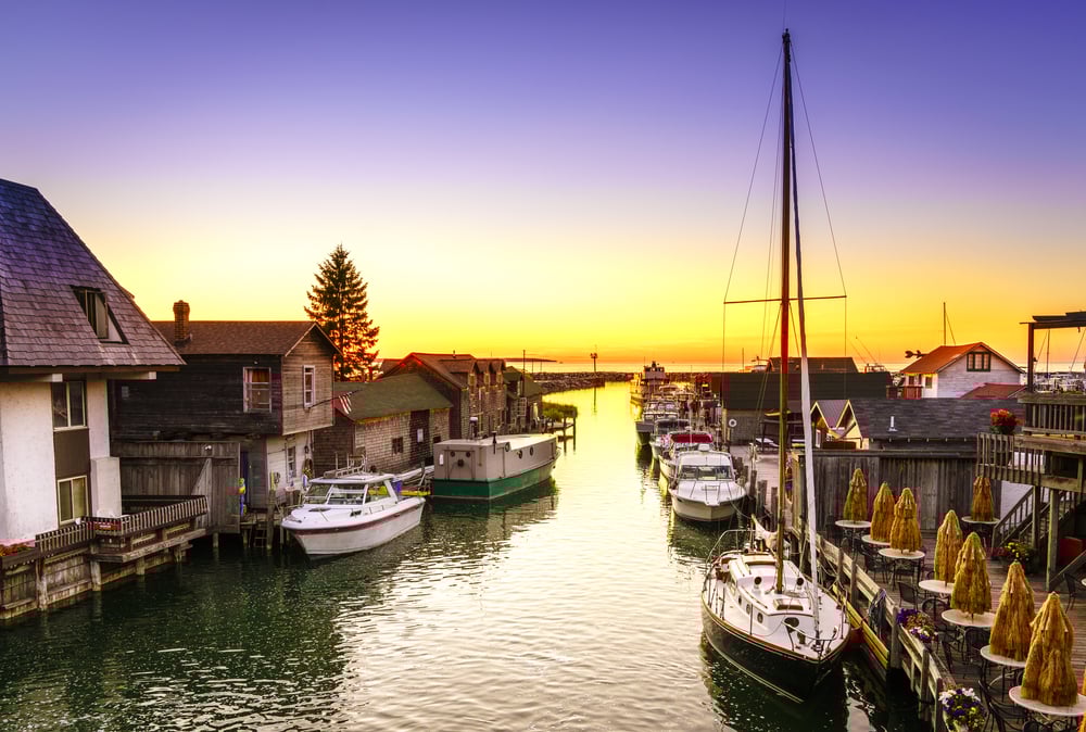 Boats docked in a narrow canal between wooden buildings at sunset, with outdoor seating on the right.