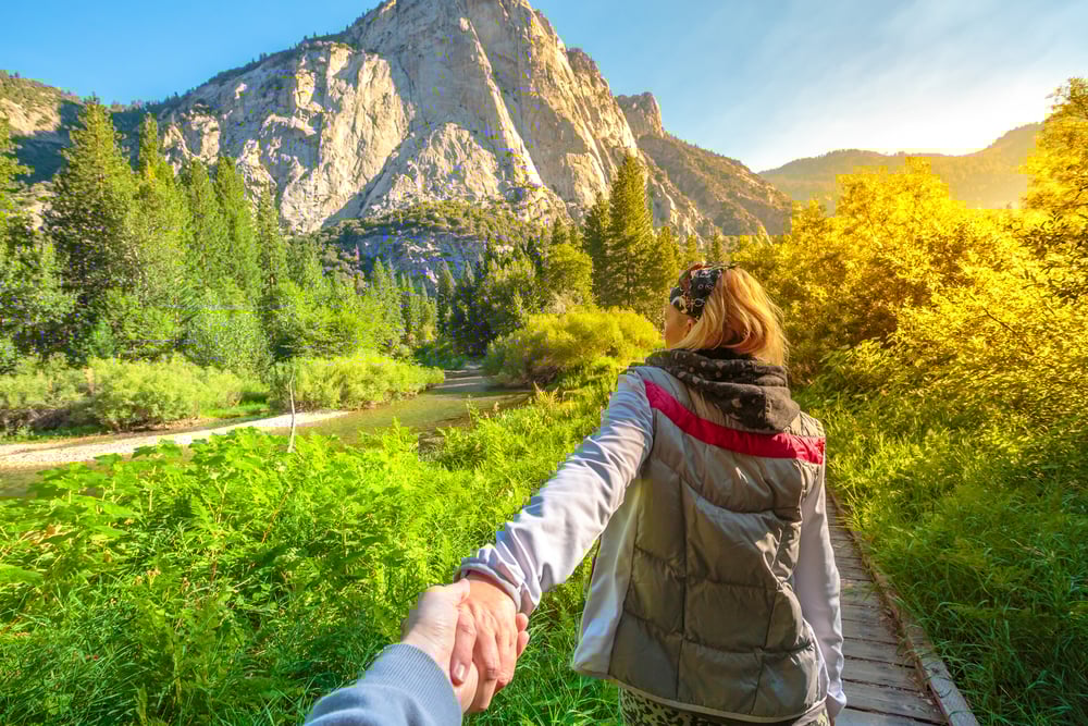 Person in a jacket leading another along a wooden path, lush greenery and mountains in background.
