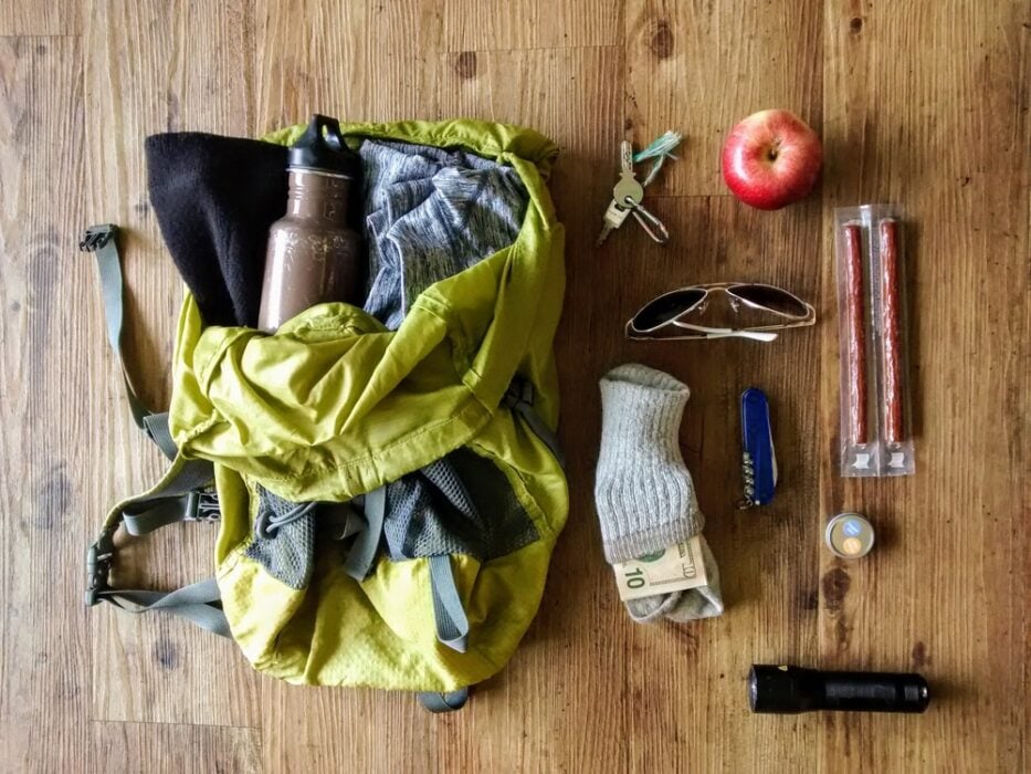 Green backpack with clothes, water bottle, keys, apple, sunglasses, sock, knife, flashlight, snacks, and lip balm.