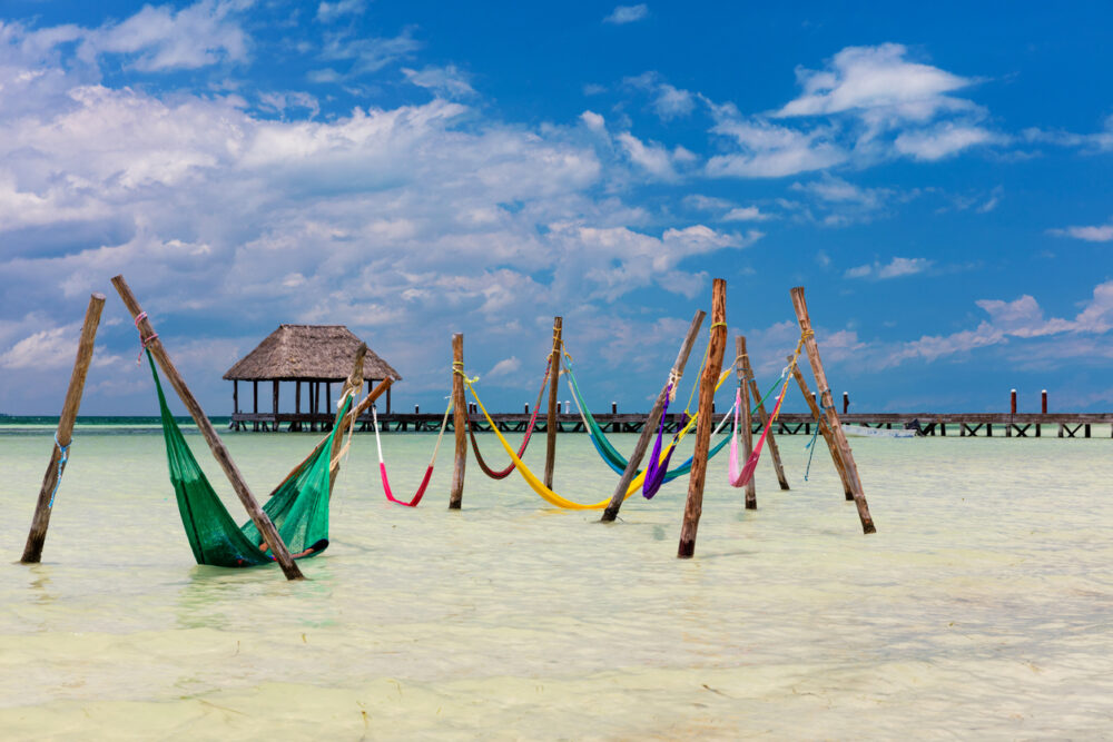 Colorful hammocks hanging over shallow water, with a thatched-roof hut and wooden pier in the background.