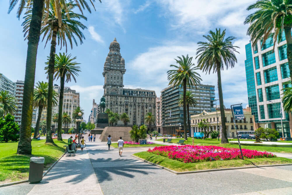 Plaza with palm trees, people walking, colorful flowers, and a tall building under a blue sky.
